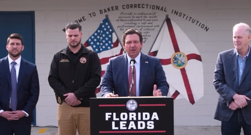 Ron DeSantis speaks at podium in front of the Baker Correctional Insitution. There are three men standing behind him. Two to his right and another on his left.