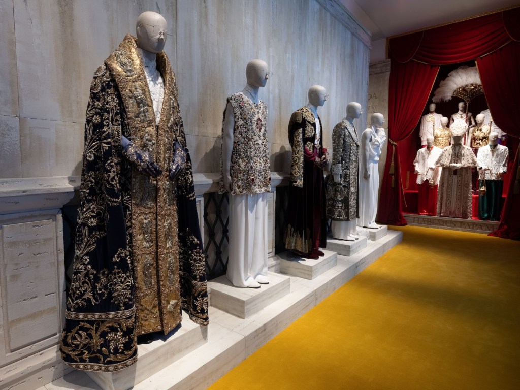 photo of mannequins donning ornate outfits in a hallway to the left and behind a red curtain in the background