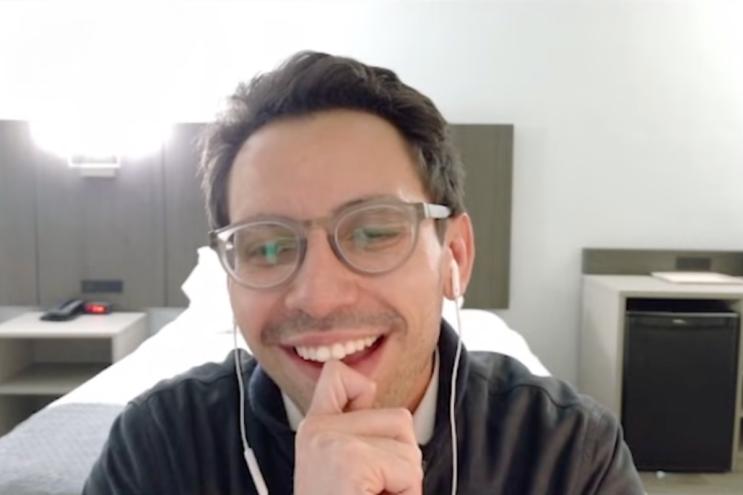 A man with brown hair wearing framed glasses and headphones, smiling at the screen