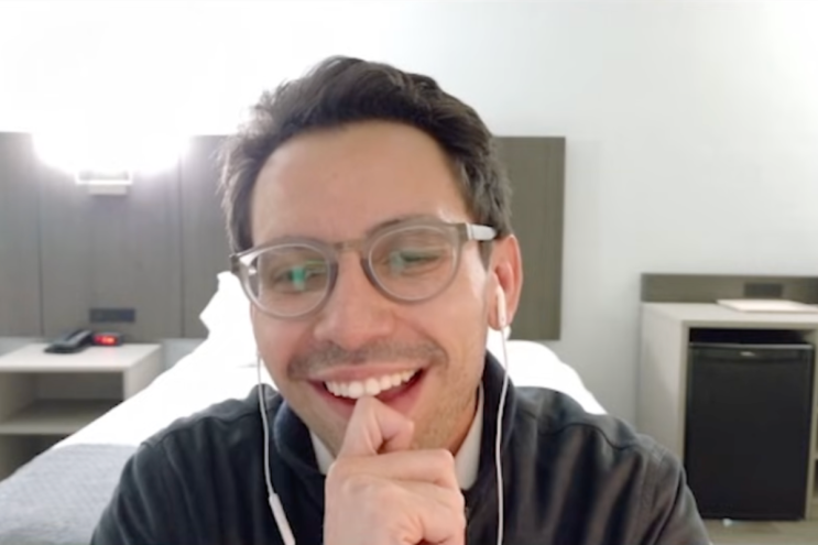 A man with brown hair wearing framed glasses and headphones, smiling at the screen