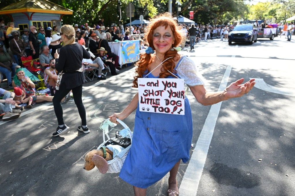 photo of a woman dressed as Dorothy from The Wizard of Oz with smudged make-up and a sign reading, "and you shot your little dog, too!"