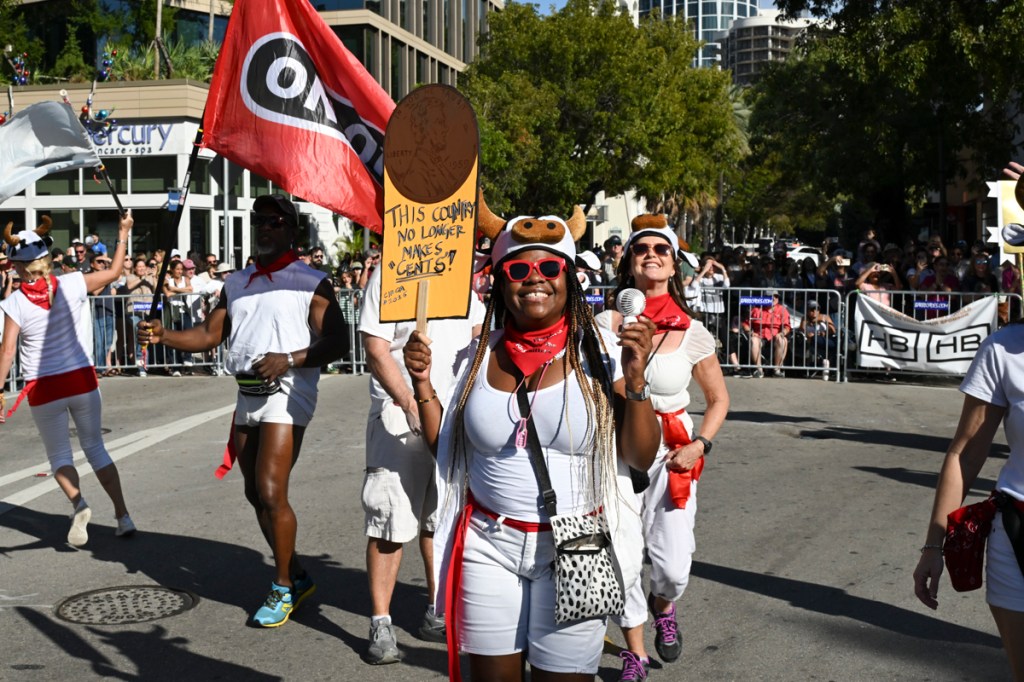 Photo of people walking in a parade. The woman in the middle holds a sign with an image of a penny on it, reading, "This country no longer makes 'cents'"