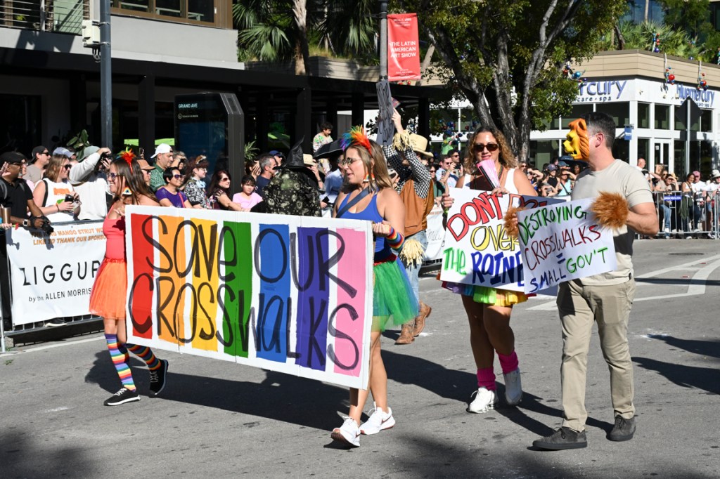 photo of people at a parade walking with a sign reading, "save our crosswalks" on rainbow-colored paint