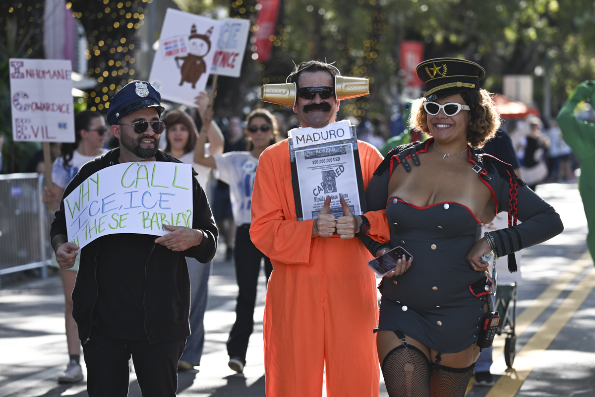 photo of three people at a parade: one is dressed as a police officer, one is dressed in a prisoner's jumpsuit holding a sign reading, "Maduro," and the other is wearing a uniform and military hat