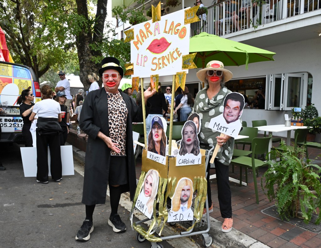 photo of two people at a booth at a parade wearing exaggerated plastic lips. The booth has a sign reading, "Mar a Lago lip service"