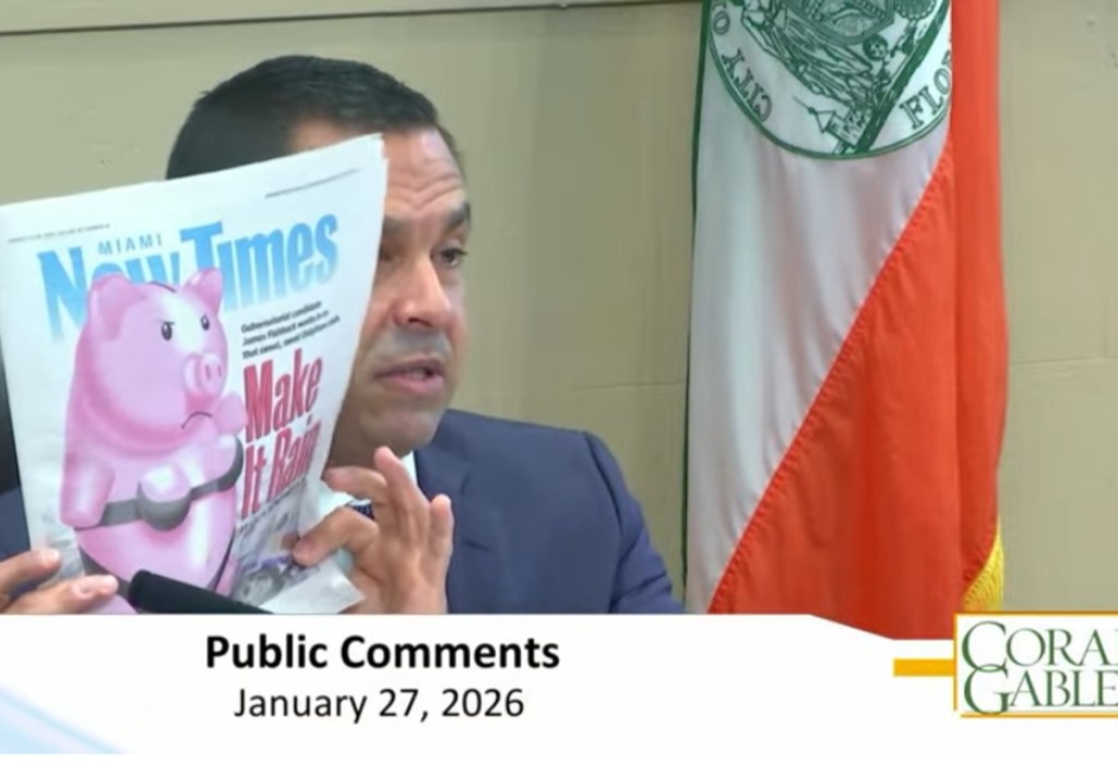 Screenshot of Coral Gables City Commission meeting and Vince Lago is holding Miami New Times cover of Piggy bank wearing a bikini. He is holding it to the right of his face.