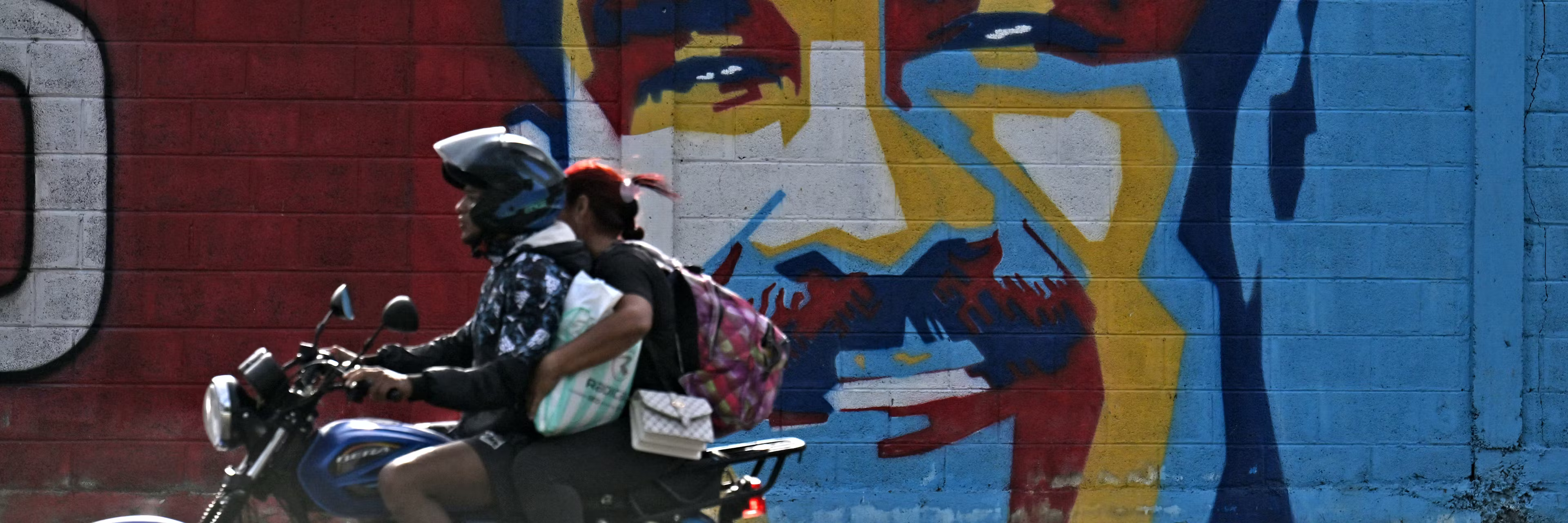Two people on a motorcycle ride past a mural of Nicolas Maduro in Venezuela