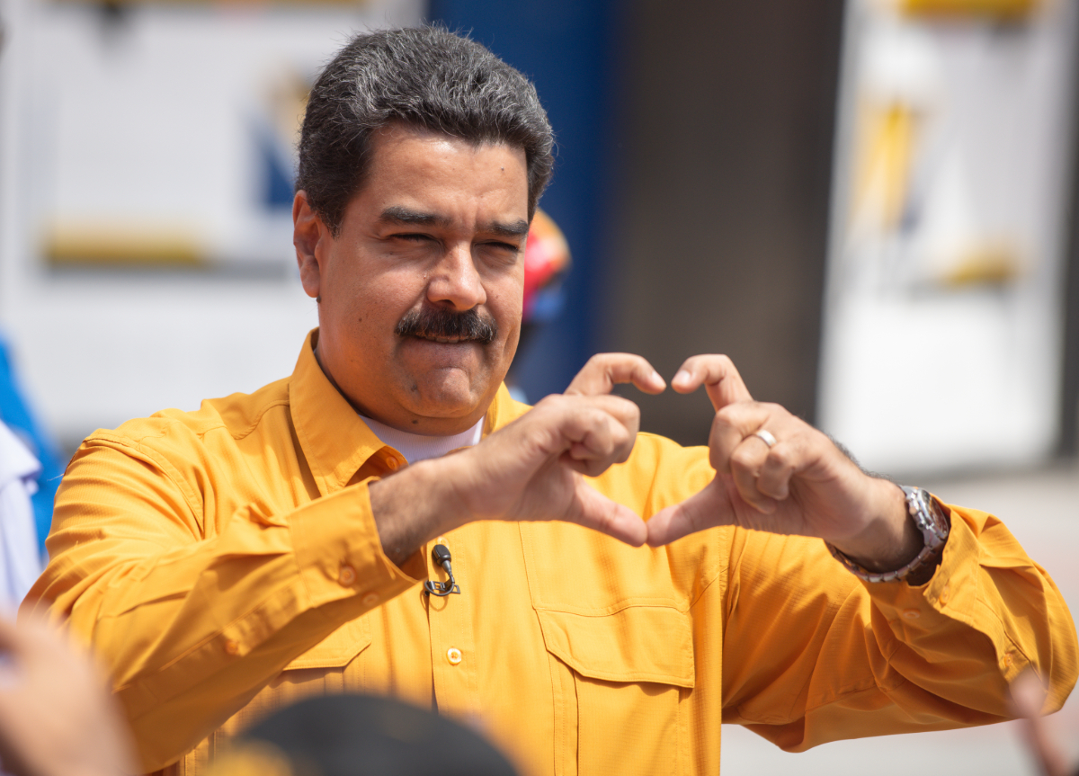 A mustachioed man wearing a yellow button down shirt makes a heart with his hands.