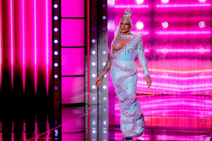 photo of a drag queen wearing a blue-and-purple iridescent gown with her left prosthetic breast exposed and covered with a pastie