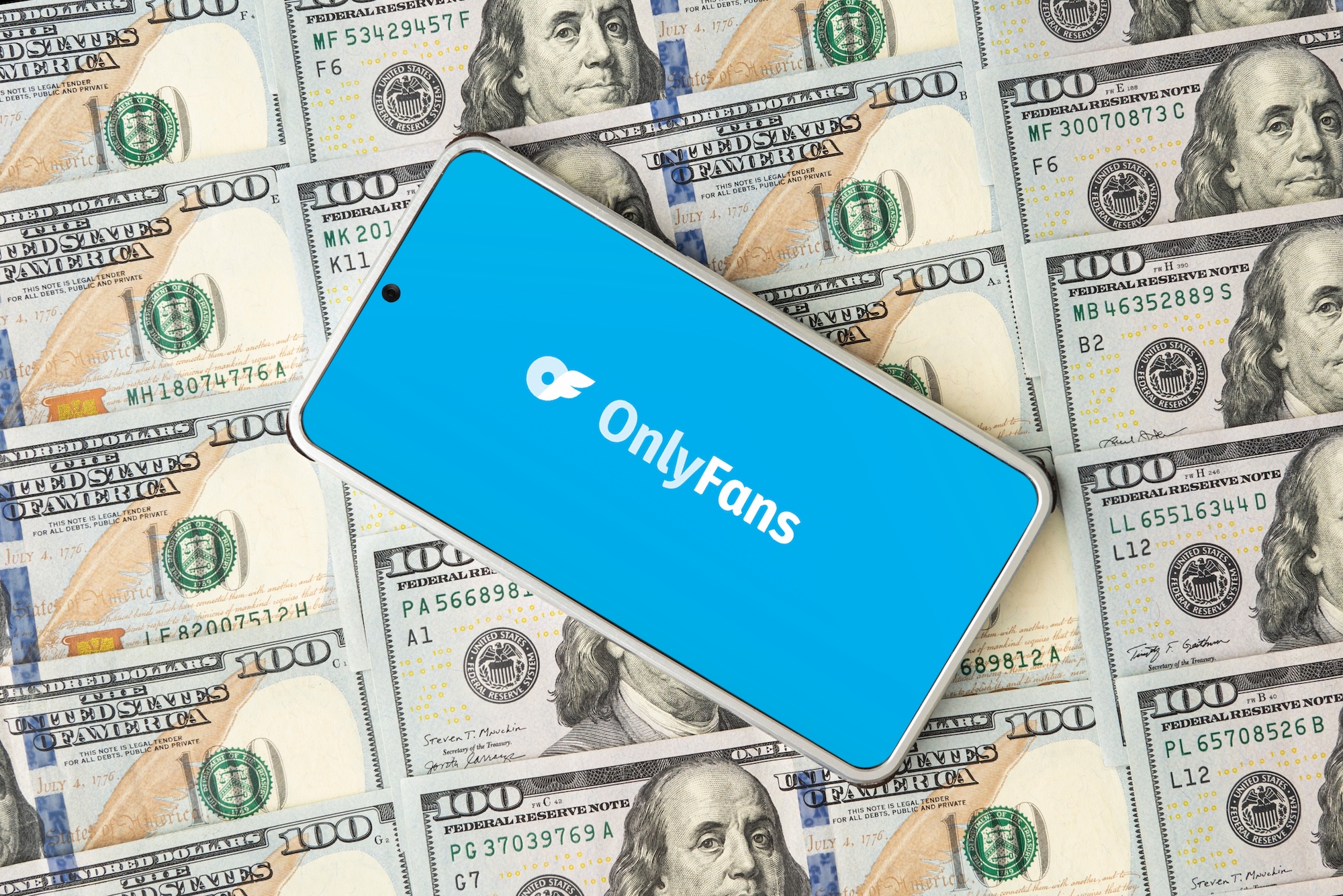 Photo of a smartphone on top of stacks of hundred-dollar bills. The phone displays the OnlyFans logo