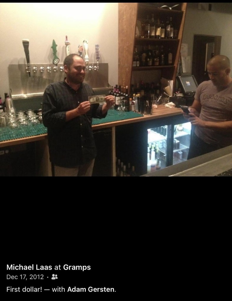 screenshot of a Facebook post showing a man in a bar holding up a dollar bill. The caption reads, "Michael Laas at Gramps. First dollar! With Adam Gersten"