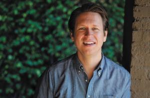 headshot of comedian Pete Holmes wearing a blue button-down shirt in front of a hedge