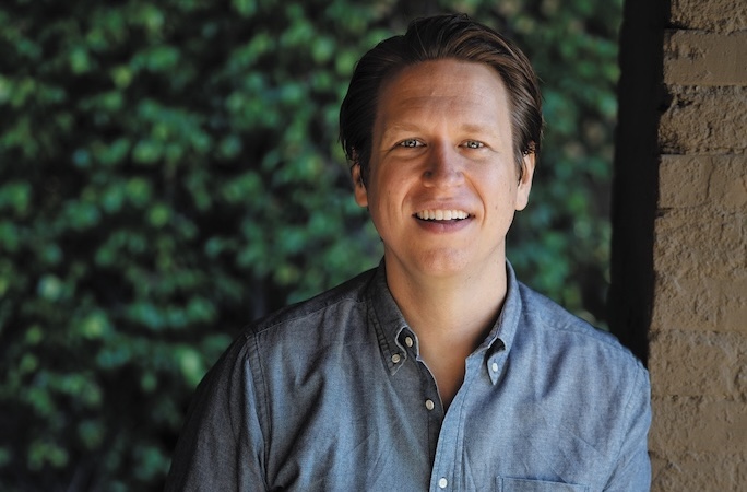 headshot of comedian Pete Holmes wearing a blue button-down shirt in front of a hedge