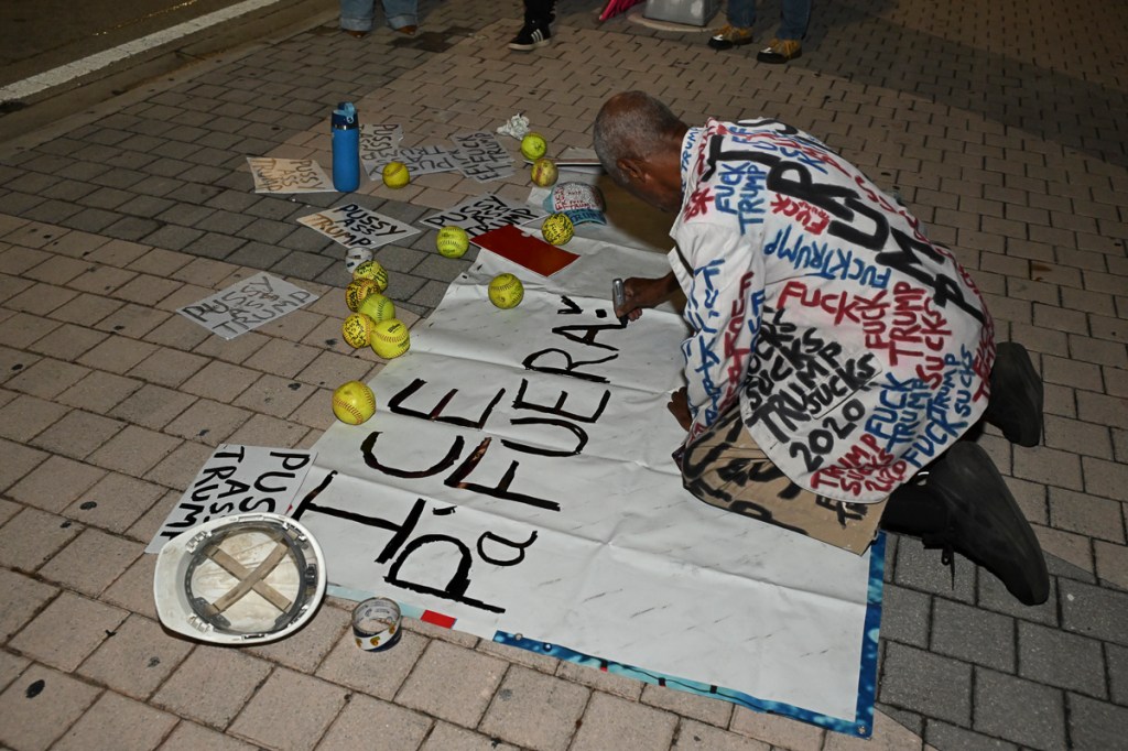 photo of a protester in a suit jacket with painted text painting on a sign on the ground