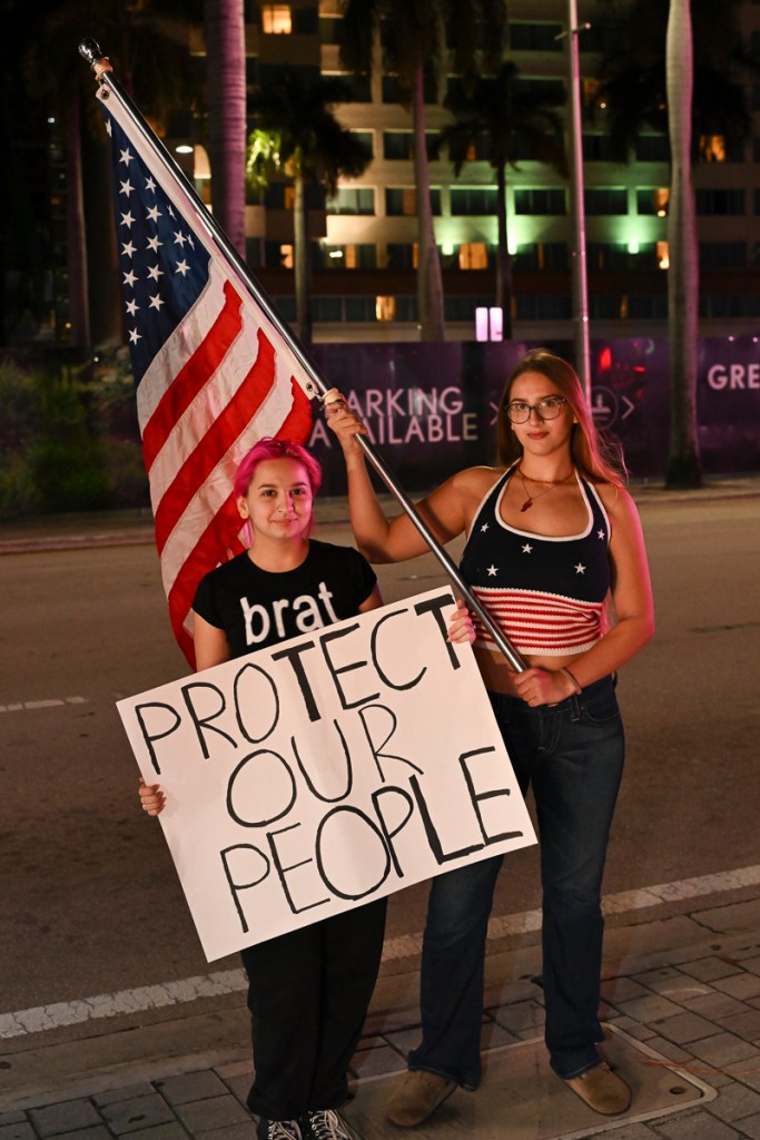 photo of two protesters holding a sign reading, "protect our people" and an American flag