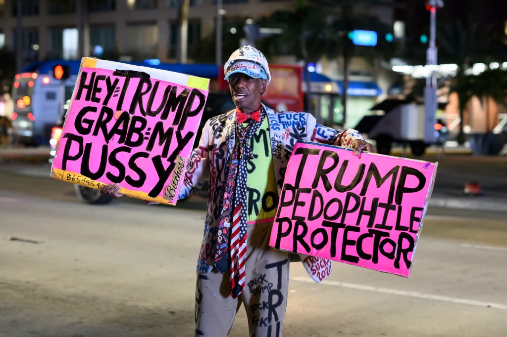 photo of a protester holding two signs. one reads, "Hey Trump grab my pussy." The other reads, "Trump pedophile protector"