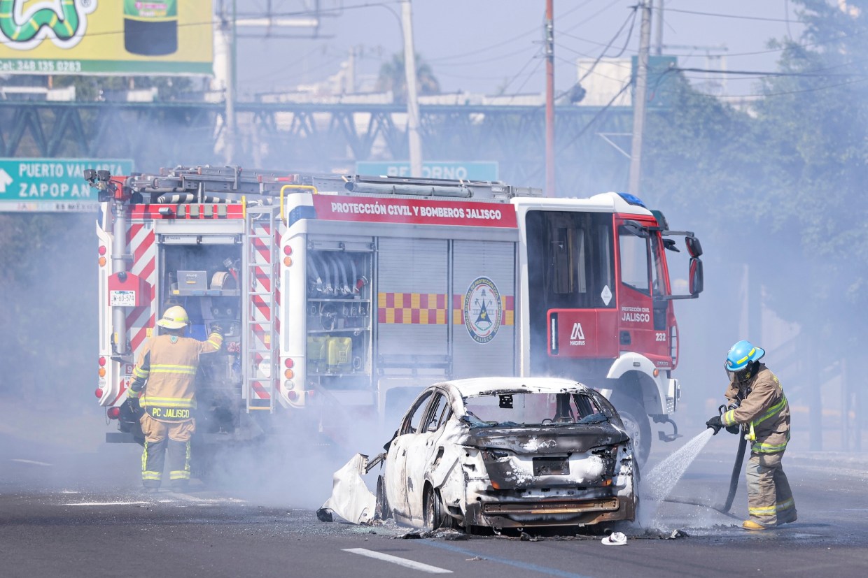 Firefighters in Mexico extinguish a fire surrounding a white car. Smoke fills the air.