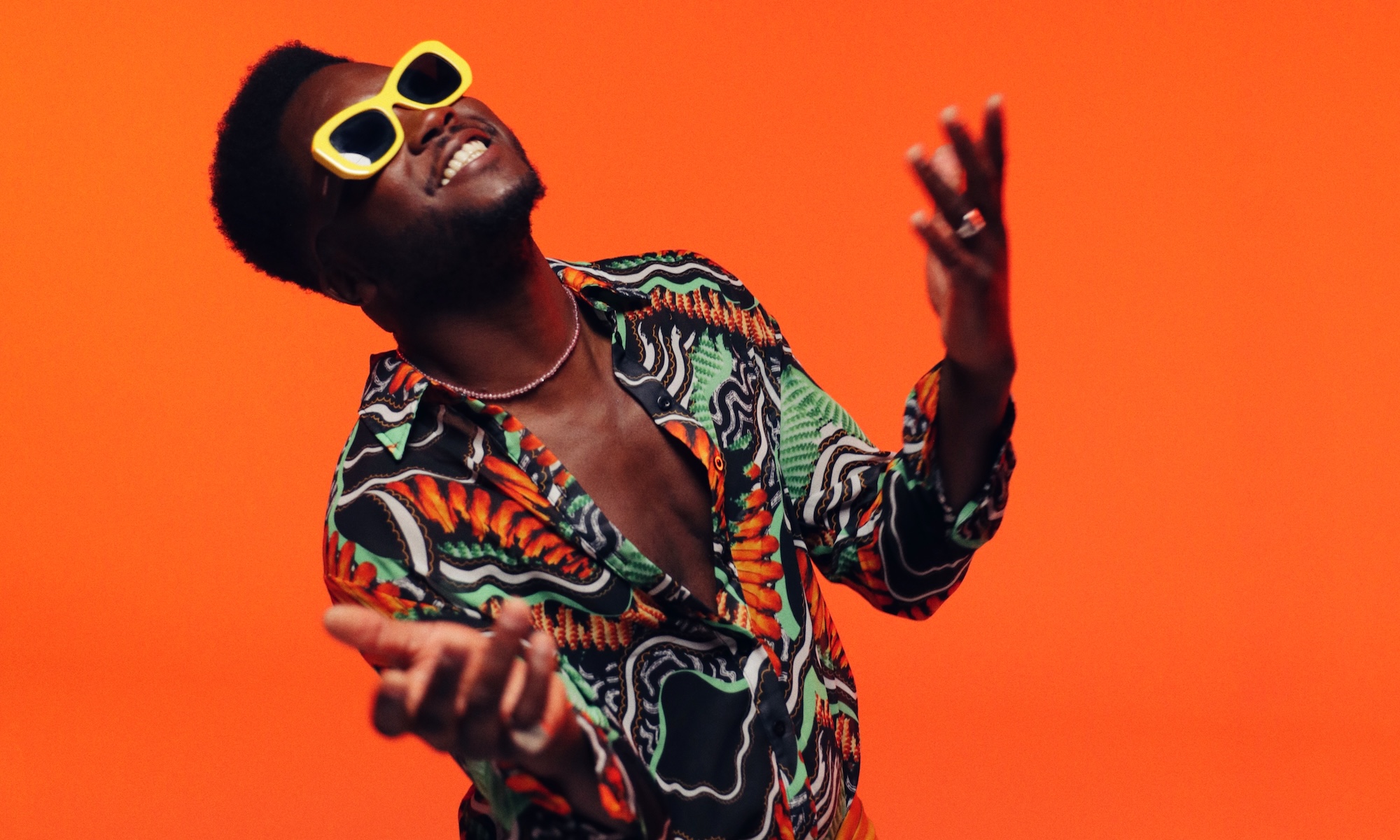 A man wearing yellow glasses and a colorful shirt against an orange background