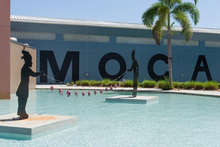 Photo of the facade of a museum with bold letters reading "MOCA." A pool is in the foreground with two black sculptures of children holding a string of fish