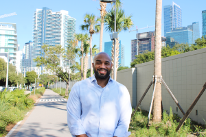 photo of a bearded Black man in a button-down shirt posing in a pedestrian pathway in with skyscrapers in the distance