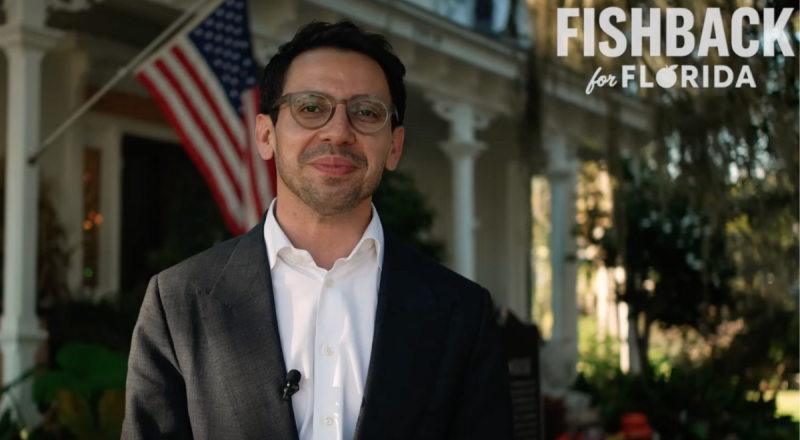 a man in a white shirt and black jacket poses in front of an American flag