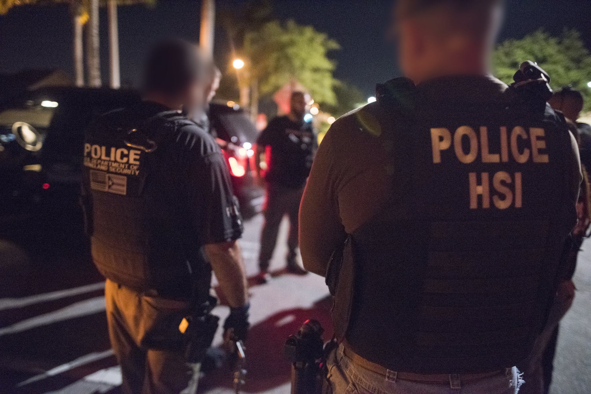 Two Department of Homeland Security agents face away from the camera outdoors at night
