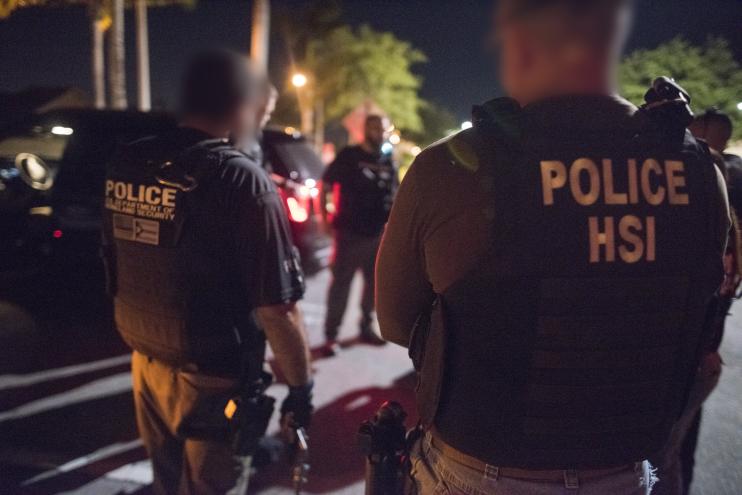 Two Department of Homeland Security agents face away from the camera outdoors at night