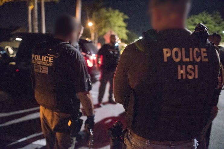 Two Department of Homeland Security agents face away from the camera outdoors at night