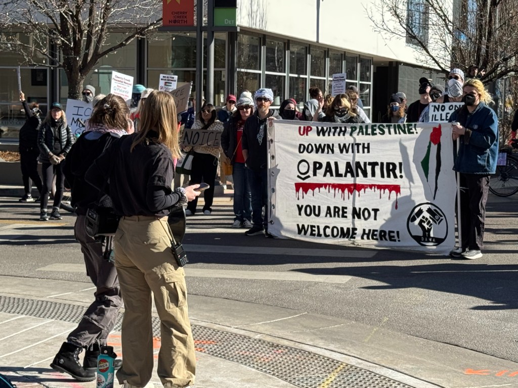 Protesters hold up a canvas sign in front of two people and that says says "Up with Palestine down with Palantir! You are not welcome here. There is a line of blood splatter under Palantir. There is also a drawing of the outline of Israel with a palestine flag drawn vertically over the outline. There is a fist at the bottom right corner.