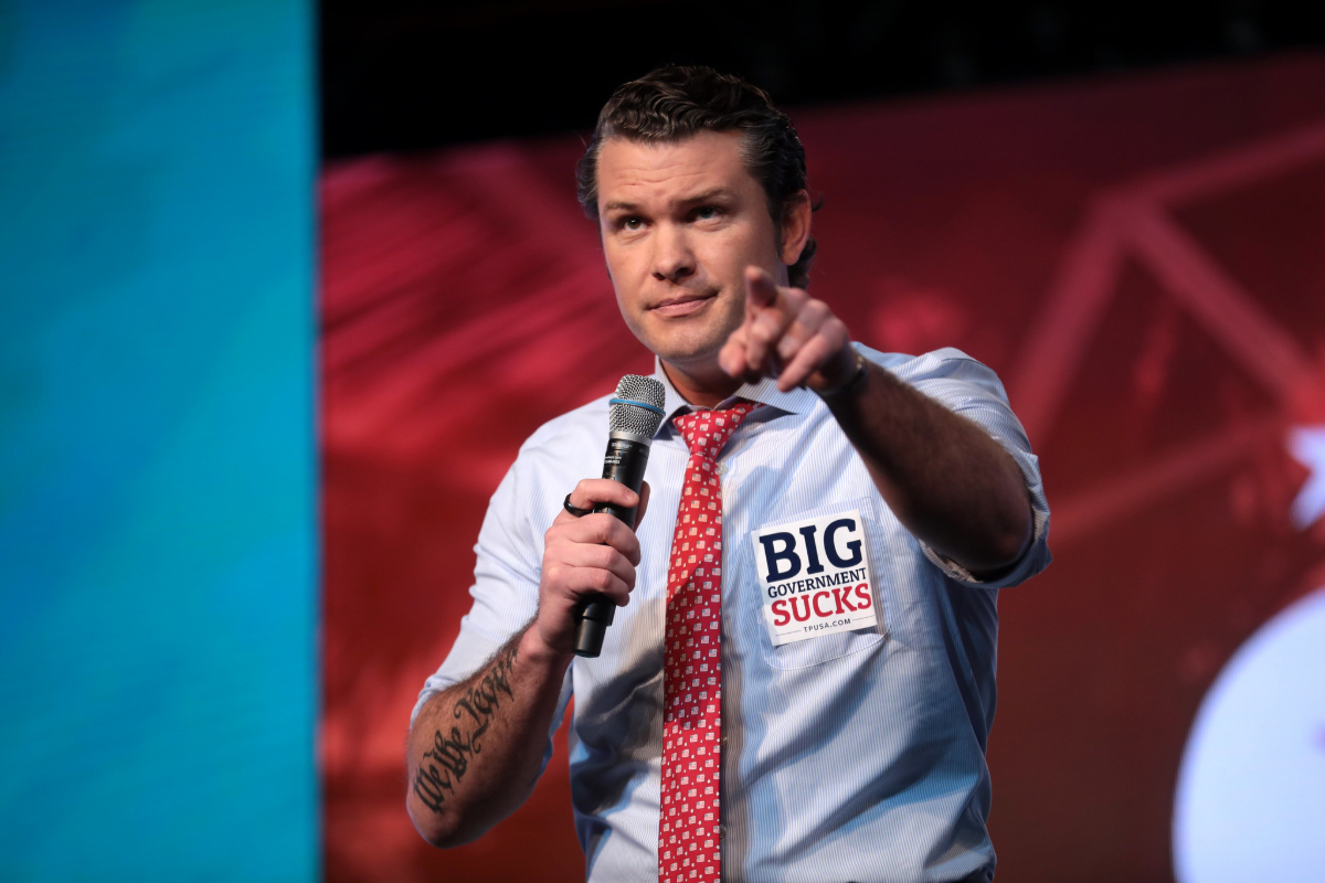 Pete Hegseth holding a microphone in his right hand and pointing with his left. He is wearing a blue button down shirt with the sleeves rolled up. He has a red tie and has a stick on the left side of his chest that says "BIG government Sucks." He has a tattoo on his right arm that says "We the People."