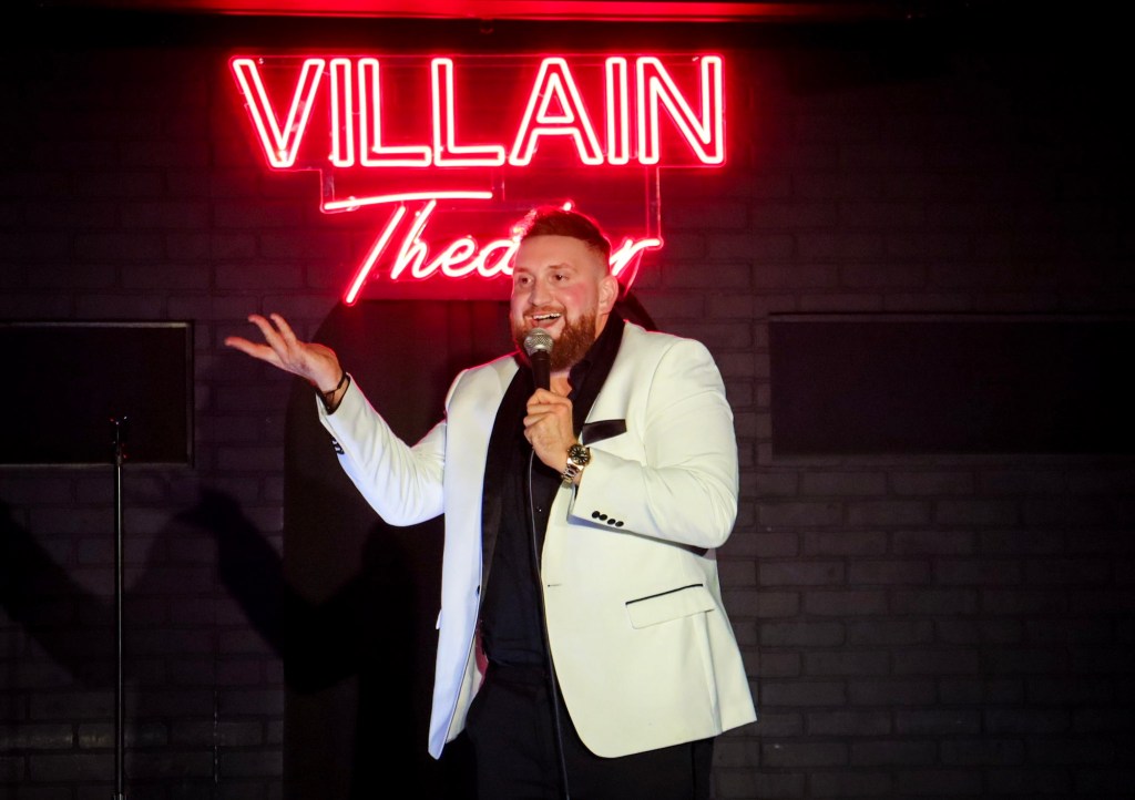 photo of a comedian in a white blazer performing in front of a neon sign reading, "Villain Theater"