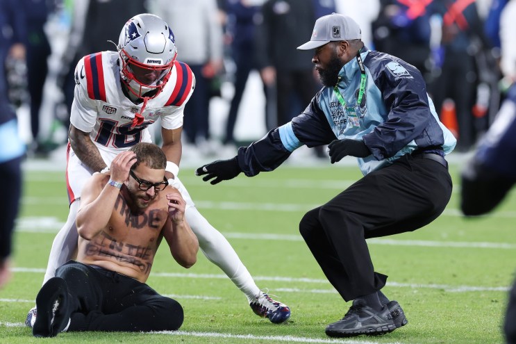 Kyle Williams of the New England Patriots and a security guard tackle a shirtless man on the field. The man is wearing black glasses and his chest says "Trade," and his stomach says "In the blindspot." He is sitting with Kyle Williams standing over him and security guard has his right arm outstretched.