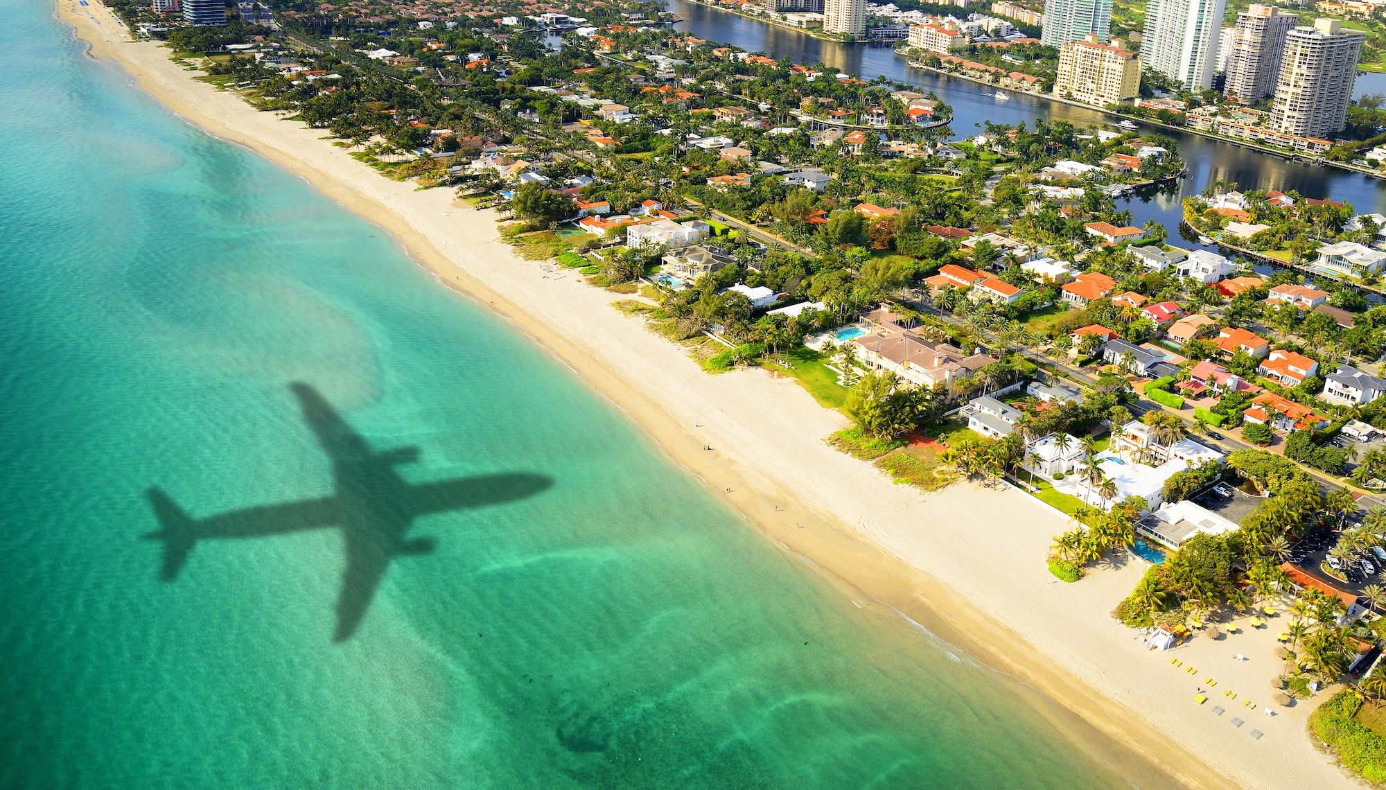 aerial photo of the shadow of a plane flying over the ocean in the direction of trees, houses, and skyscrapers in Miami