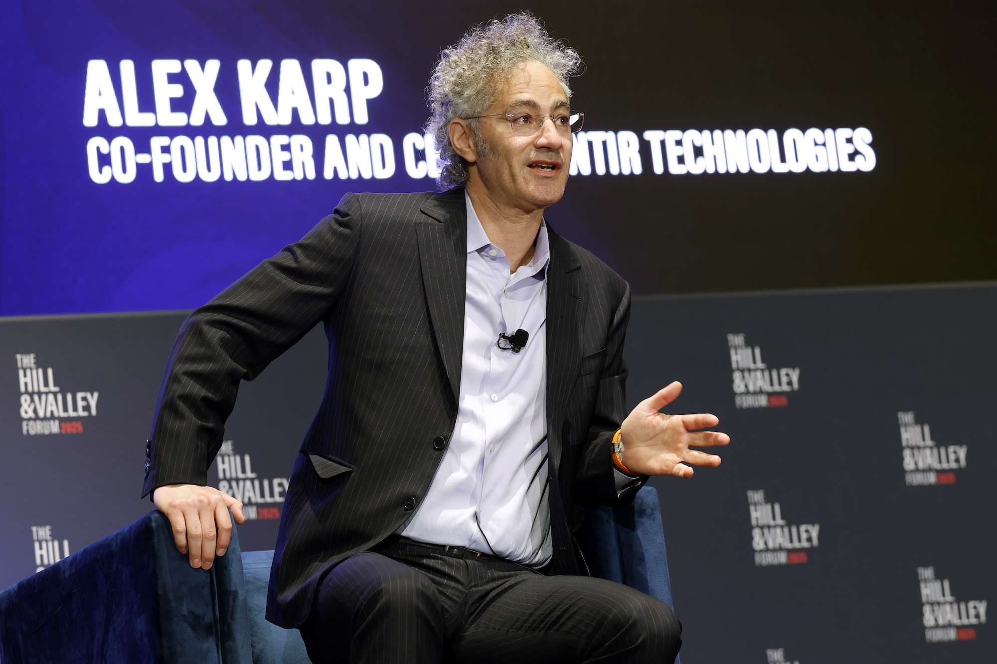 Alex Karp slightly sitting out of his chair while speaking at a panel
