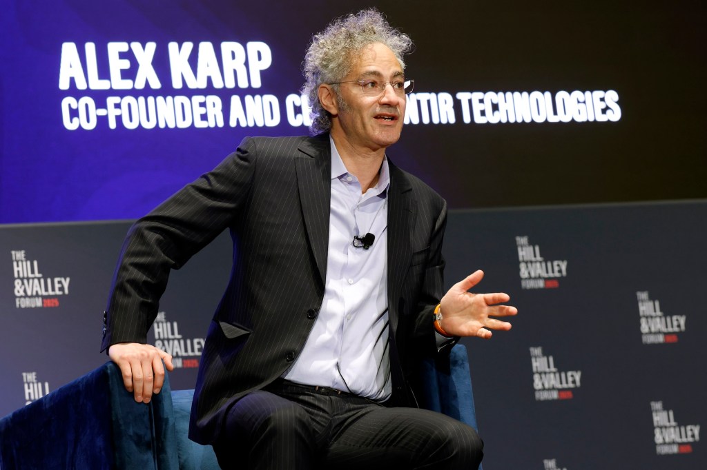 Alex Karp slightly sitting out of his chair while speaking at a panel