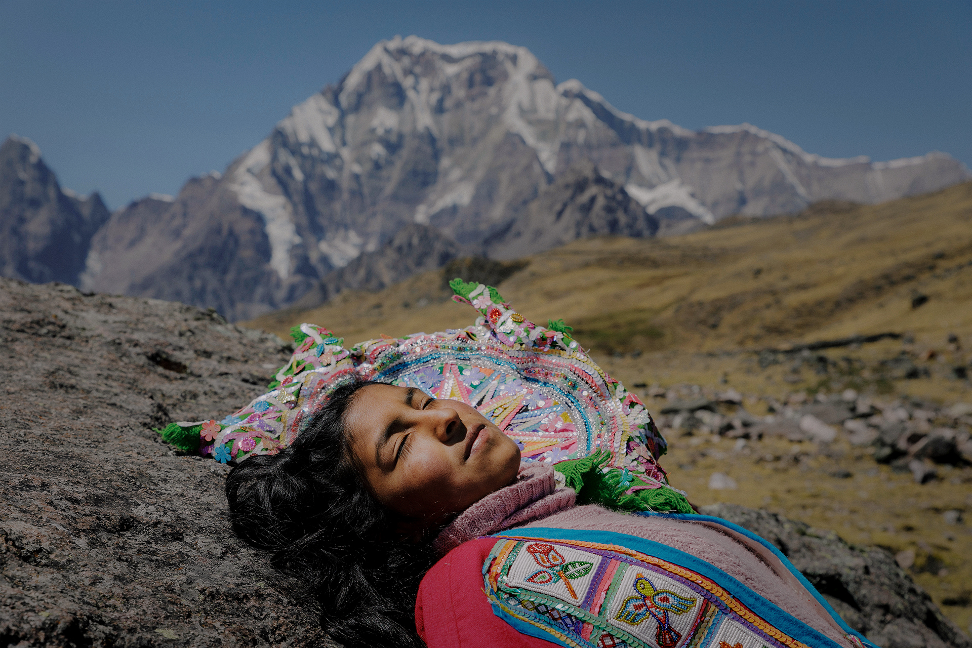 Photo of an Indigenous woman in colorful traditional clothing lying on the ground in a mountainous area. Behind her are mountains in the distance