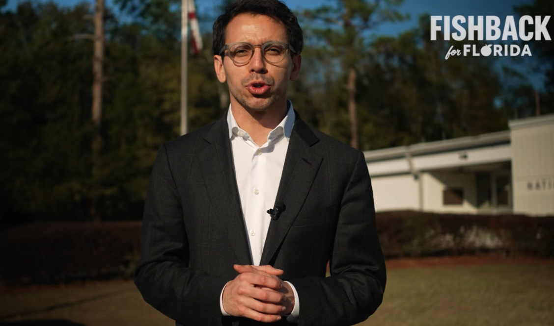 A photo of a bespectacled man wearing a suit and speaking to a camera outdoors near a one-story house.