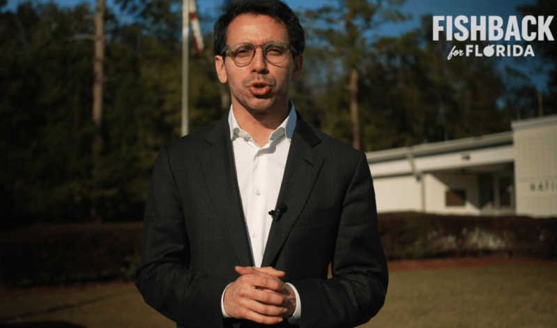 A photo of a bespectacled man wearing a suit and speaking to a camera outdoors near a one-story house.