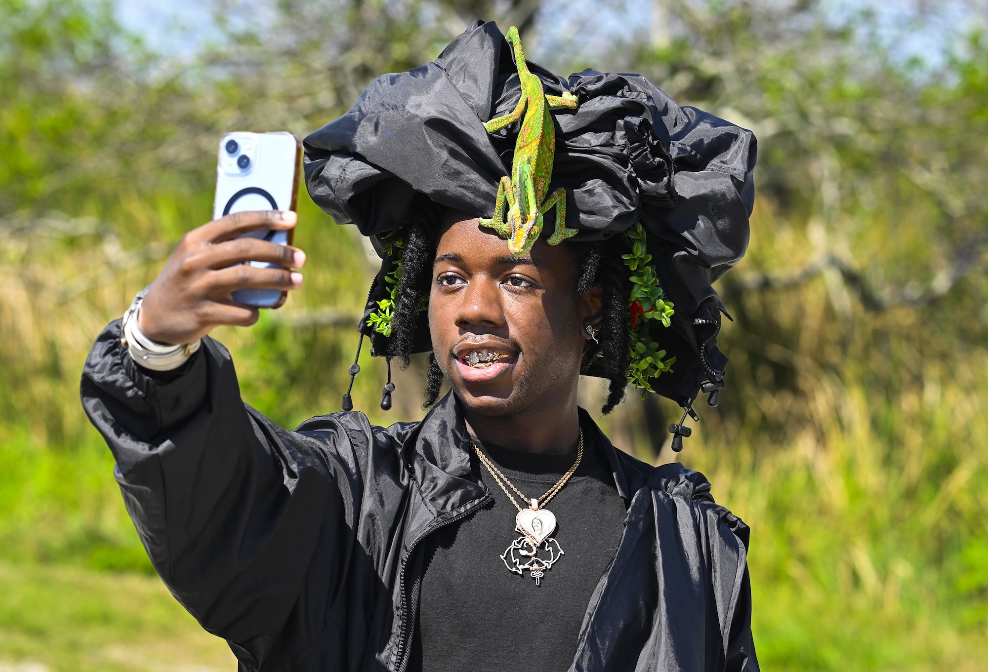 Photo of a young Black man with a jacket on his head and a chameleon on top of the jacket. He takes a selfie on his cell phone