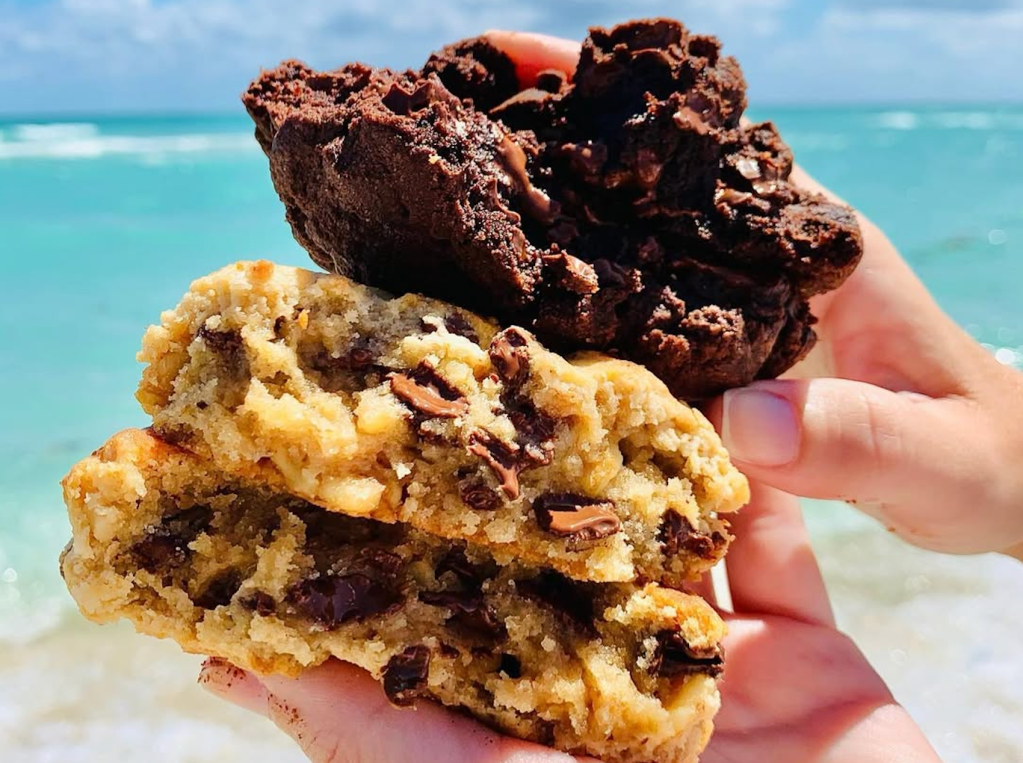 two Levain chocolate chip cookies being help at the beach in the sun and in front of the ocean in Miami