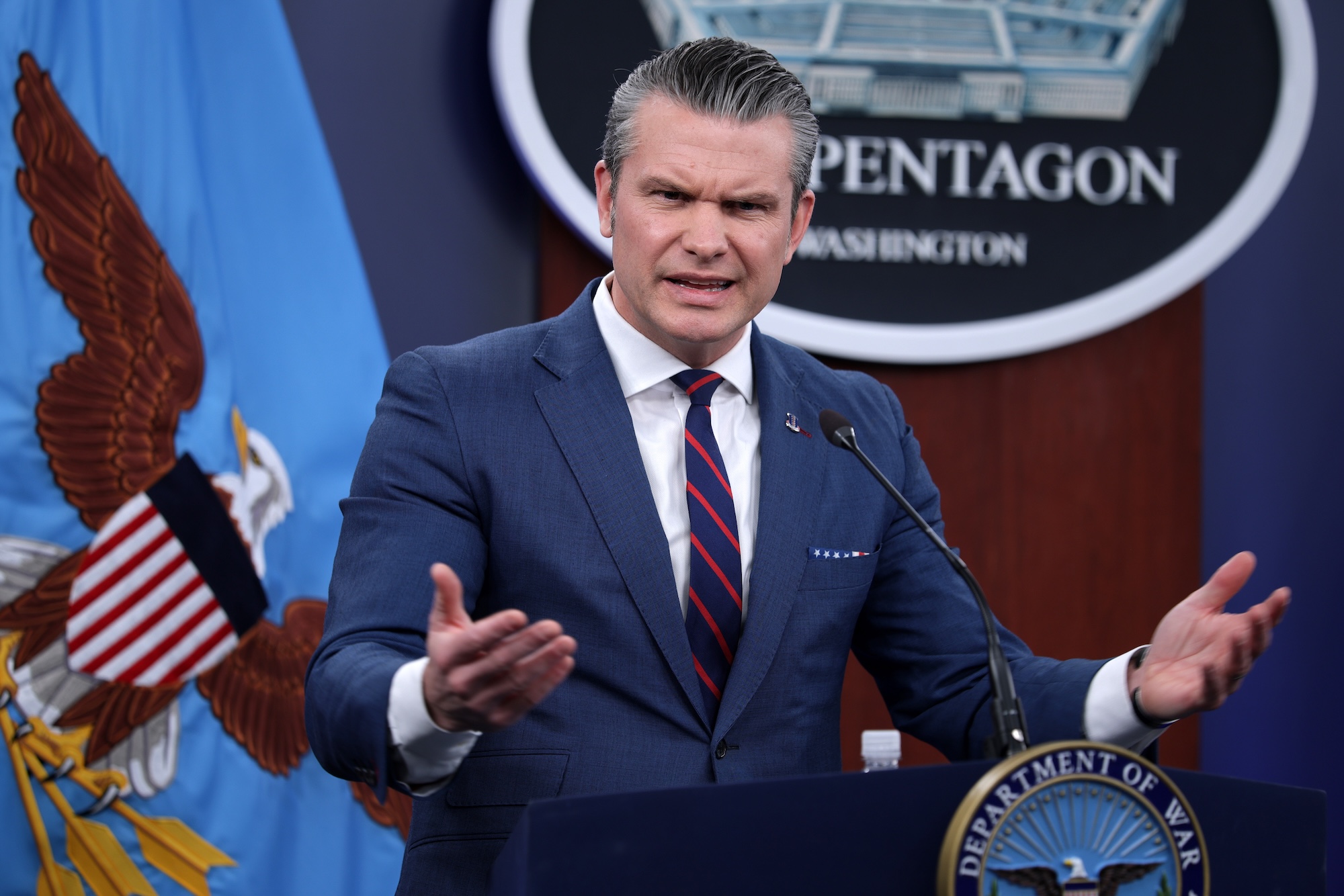 Pete Hegseth speaks from the Pentagon with his hands out. He is wearing a blue suit with a blue tie with red diagonal stripes.
