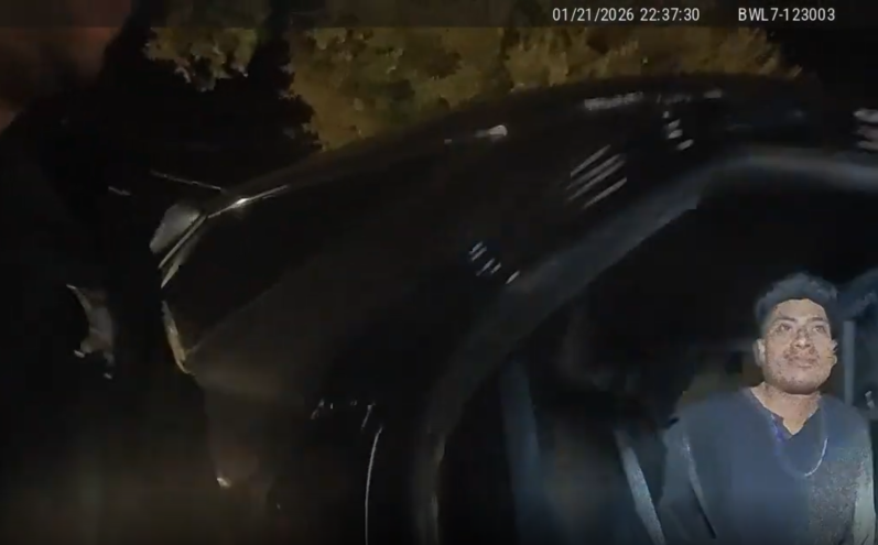 Screenshot from police body cam video shows a young man sitting inside a patrol car with a flashlight being held on his face.