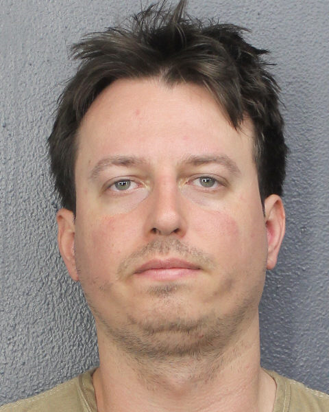 A white man with messy black hair and stubble poses for a mugshot with a blank expression