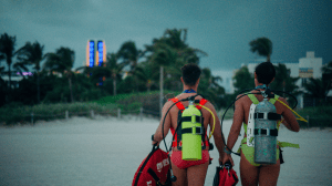 photo of two men wearing Speedos and oxygen tanks walking on sandy shores in the direction of the city and palm trees