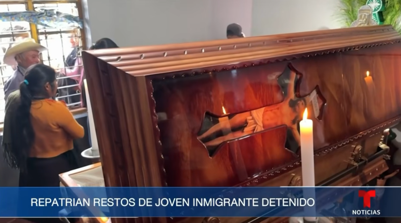A screenshot of a TV broadcast showing a candle lit in front of a wooden casket at a burial.