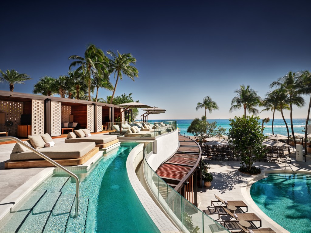 photo of a hotel pool area showing a pool with daybeds on a top floor overlooking a pool and tables with umbrellas in front of the ocean