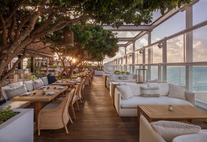 Photo of outdoor seating at a hotel restaurant showing tbles with booths and wicker chairs on the left and couches and coffee tables on the right. The terrace is enclosed in glass and the ocean is visible on the right