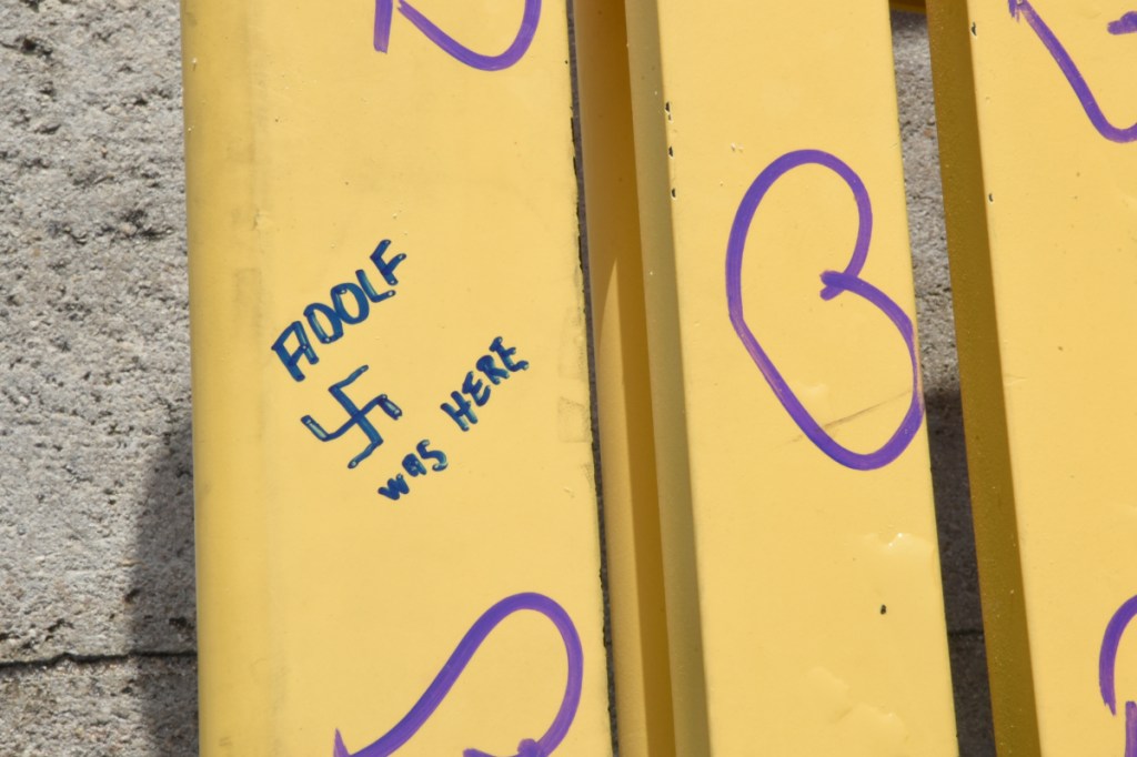 A photo of a yellow bench scrawled with the phrase "ADOLF WAS HERE" and a swastika symbol.