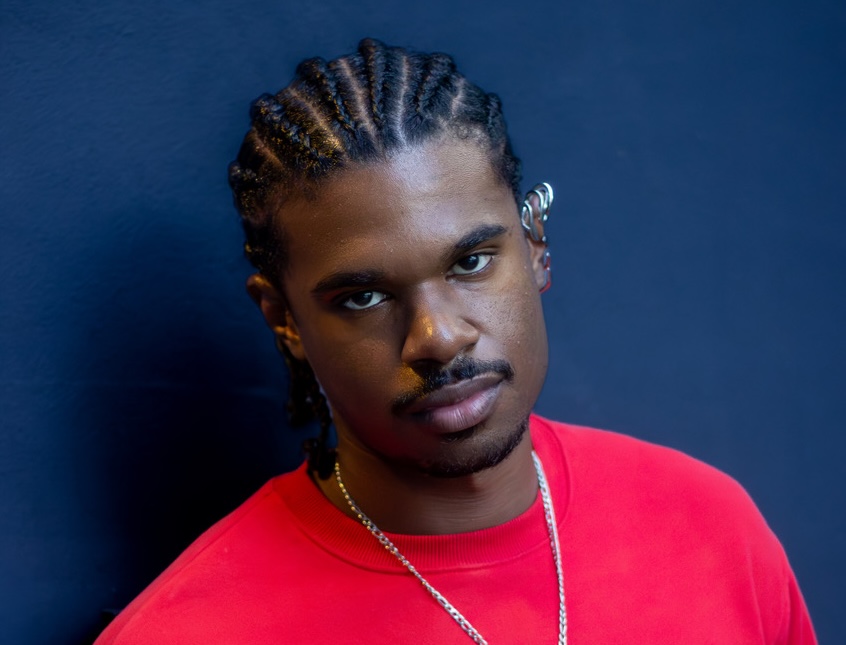 Photo of a young man with braids looking at the camera wearing a red sweater.