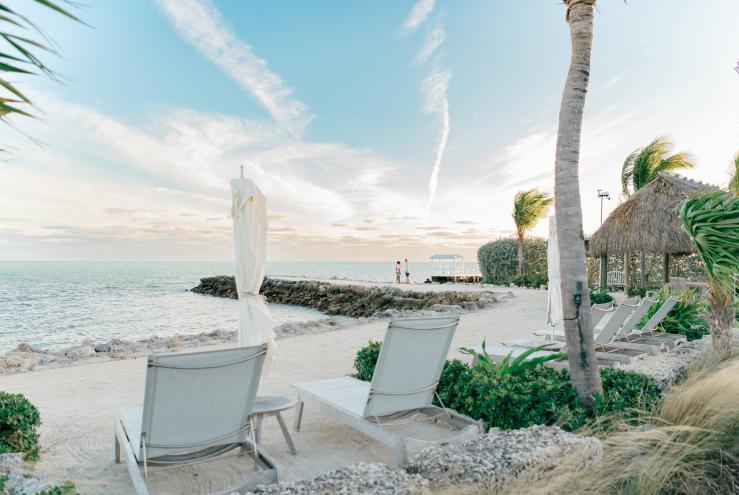 photo of a beach showing two beach chairs and folded beach umbrellas next to a tiki hut with two small people on a jetty in the distance
